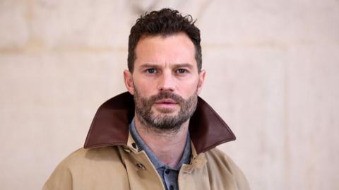 Jamie Dornan looks into the camera. He has dark hair and a short beard. He is wearing a beige coat with a brown collar with a blue shirt underneath.