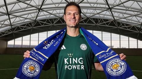 Goalkeeper Asmir Begovic smiling wearing a green Leicester City training top and wearing a Leicester City blue scarf which he is holding round his neck. 