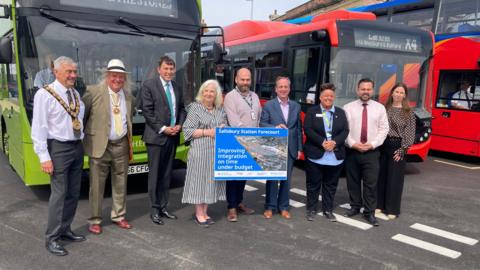 Nine people dressed smartly stand in a line in front of a series of buses at Salisbury Station forecourt.