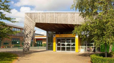 An entrance to a school building. Columns are painted yellow around a glass door, with a large wood pillar and roof, in front and above it respectively. Other buildings can be seen on the left and there is a tree on both the left and right of the photo. The words 'The Willows' are written above the door.
