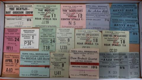 17 pastel-coloured ticket stubs laid out in a cardboard box for bands including The Beatles, The Searchers and Cliff Richard, all priced in shillings and pence.