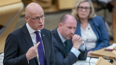 John Swinney, dressed in a dark suit and purple tie, answering questions in the Holyrood chamber