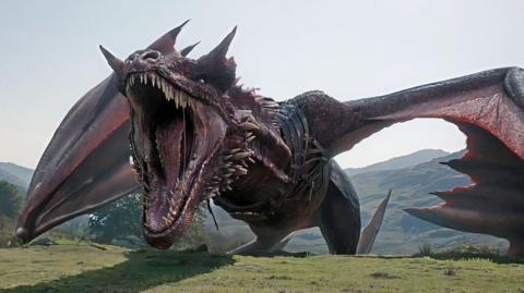 CGI of a giant red-black dragon, open mouthed, its wings out-stretched as it appears to stride rather than fly atop a mountainous landscape
