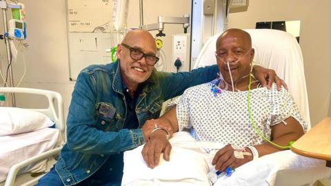 Garry Christian, smiling wearing black glasses and a denim jacket, clasps the arm of Lionel Duke, who is also smiling while sitting up in a hospital chair, wearing a patient's gown, with various tubes attached to parts of his body. 