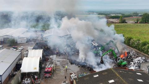 Two green JCB diggers at a scrapyard, and a fire engine. There is smoke billowing from the site.