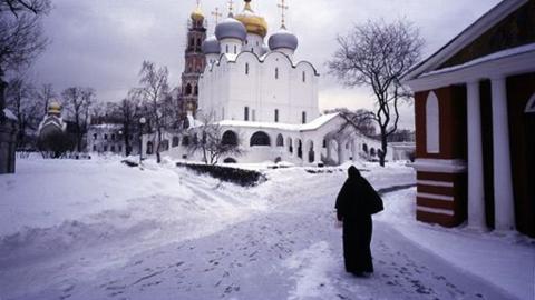 A person walks through a snowy street in Russia. There is a white building in the background