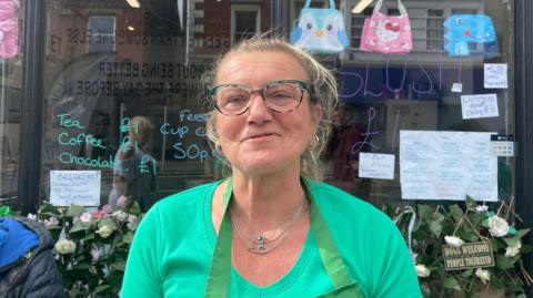 Sam Elliott has blonde-brown hair tied back in a ponytail. She is wearing a green t-shirt green apron and green glasses and smiling. She's standing in front of a cafe window. Menu items and prices are displayed on the window in colourful writing. 