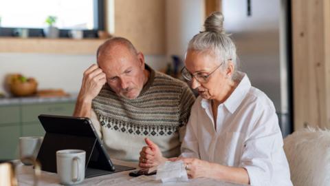 A senior man and woman sit at a table with a tablet device and look at receipts