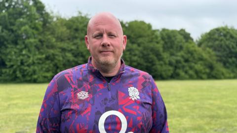 A man is standing in a field with trees behind him. He is wearing a pink and purple patterned rugby top. He is bald with a short beard.
