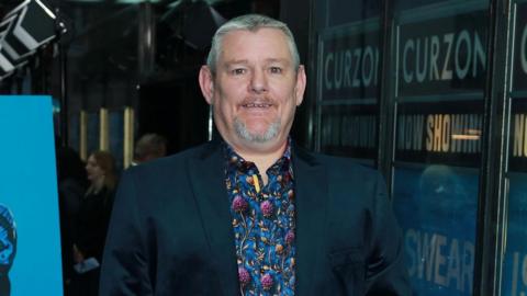 John Davidson has short grey hair and neatly trimmed beard and mustache. He is wearing a dark blue jacket over a colourful shirt with the colours blue and purple. Mr Davidson is standing in a cinema and behind him is a poster for I Swear, a film about his life.