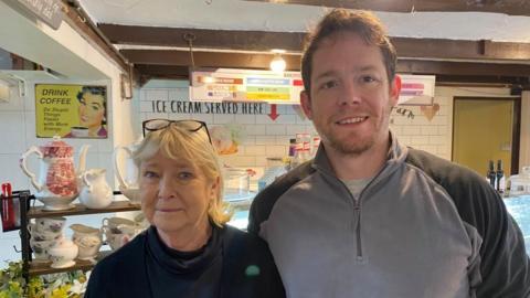 A man and a woman stand side-by-side in front of a cafe counter. The man is on the right wearing a grey quarter-zip jumper with black shoulders. The woman is in a black jumper and has her glasses resting on her head.