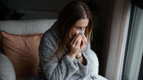 A woman with long brown hair in a grey dressing gown blows her nose