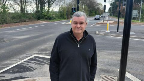 Marcus Kravis is standing on the renovated crossing in Dunster. He is wearing a black fleece and is smiling at the camera.