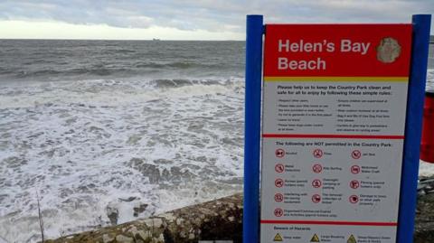 The image shows waves at the edge of a concrete pathway at Helen's Bay beach. The sign in the foreground is red and lists do's and don't's for the country park. 