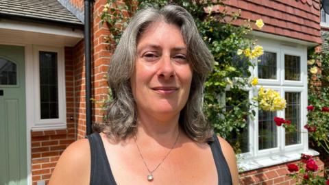 Jessica is standing in front of her house - a red-brick property with white windows, a sage green door, and red and yellow roses growing up the walls. She is wearing a black vest top, and is looking into the camera and smiling slightly. 