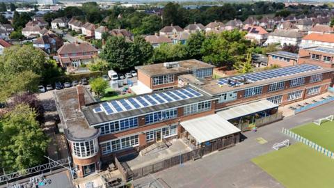 An aerial drone shot shows the roof of the school, covered in solar panels. Some workmen can be seen working at some of the panels on the roof. 