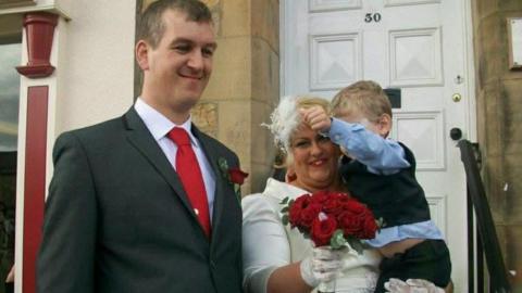 Joanne Howie is wearing a long-sleeved wedding dress with white gloves. She is holding a bouquet of red roses and a little boy. Her husband Christina Howie is standing next to her outside the white door of the registration office. He is wearing a red tie and a dark grey suit. He has short brown hair.