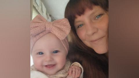 A selfie of a smiling mum with long brown hair with a fringe, and her baby daughter, who is smiling and has a large pink bow on her head.