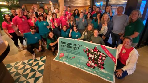 A group of people indoors pose together holding a large banner that reads 'Are you up for a challenge?' Some are wearing red shirts, others teal. The banner features logos for 'CoppaFeel!', 'boob or bust', 'MONDO', and 'dish', along with an illustration of a mountain with flags. In the background, there are signs labeled 'Kitchen' and 'Exit'.