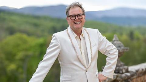 Alan Carr, a man with unkempt hair and thick-rimmed glasses. He is standing with one hand on his hip and the other leaning on a wall in front of lots of greenery. He is smiling which shows off a gap in his teeth. He is wearing a cream suit jacket with gold beading on it.