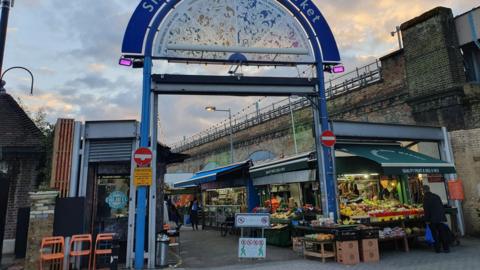 A blue metal arched entrance, an stalls on either side. A fruit and vegetables stall is seen on the front right corner, and a falafel stand on the front left corner. 