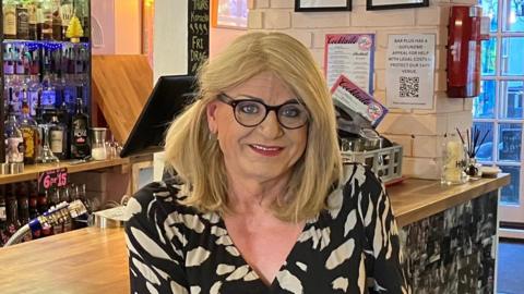 A woman sitting on a stall in front of a bar. She has blonde hair, glasses and is wearing a black and white dress.