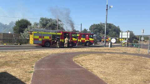 Fire engines parked up along a road with smoke rising in the background