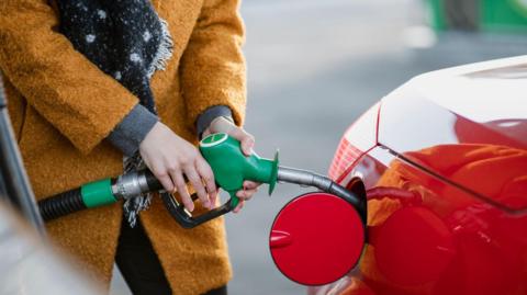 Close up shot of a young woman wearing an orange coat filling her red car with fuel.