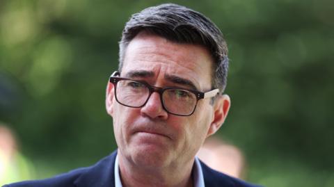 A middle aged man with dark, slightly greying hair. He is wearing a dark blue suit jacket, a white shirt and dark , squarish rimmed glasses. His lips are pursed and he has a serious look.
