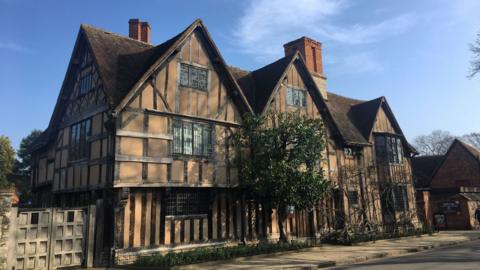 A timber‑framed house with steep roofs on a street under a clear blue sky.
