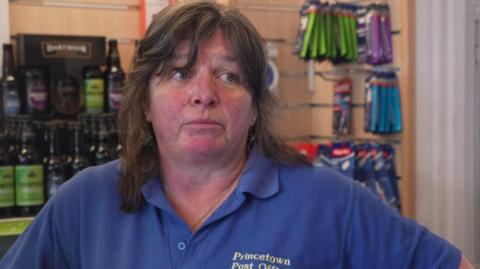 Jane Liversidge has shoulder-length dark brown hair. She is wearing a blue polo shirt with the words Princetown Post Office and Store embroidered.