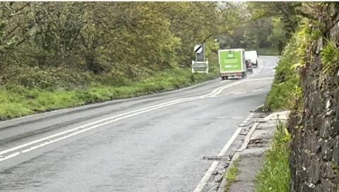 A country lane bordered by green verges with a group of vehicles in the distance