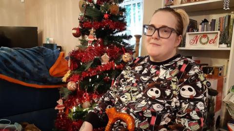 Charlotte Hale is sitting in her living room with a bookshelf behind her, which has a decorative sign saying Noel, and there is a Christmas tree with red tinsel and baubles. Miss Hale is wearing glasses and holding a walking stick.