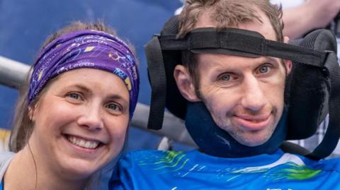 A woman with long brown hair tied back smiles on the left of the frame while a man in a wheelchair, his head supported by a strap across his forehead, smiles from the right of the frame