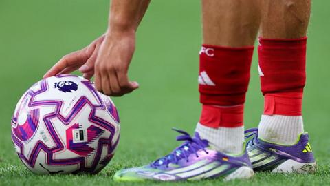 Image of a footballers two feet, with with a hand reaching down to touch a ball placed in front of him.