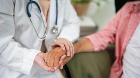 A doctor holds a woman's hand in a consultation. The doctor has a white coat and a stethoscope.