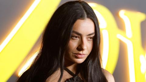 Charli XCX looks just off camera with a serious expression. She has long dark hair down past her shoulders and behind are illegible large letters in neon yellow lights.