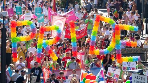 Pride Cymru supporters parade through the city centre in Cardiff. Rainbow balloons spell PRIDE and attendees line the streets.
