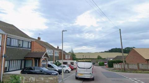 A street of houses with cars and a van parked in front. In the distance are green fields.  