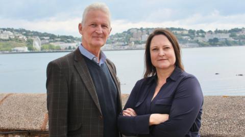 Mark Kingscote and Alison Hernandez are smiling at the camera as they stand in front of a harbour wall with the sea behind them and houses visible on land in the distance. He is wearing a checked jacket over a blue jumper and shirt and she is wearing a navy blouse and has her arms crossed.