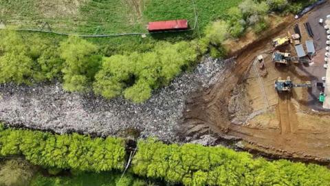 Machinery on an area of brown earth next to a large mound of flytipped waste between two rows of green trees.