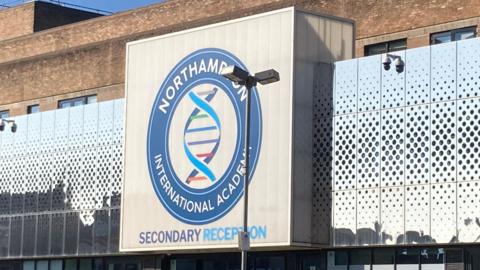 A close-up photo of the Northampton International Academy logo - a multi-coloured helix in a blue circle - on the front of the school building. 