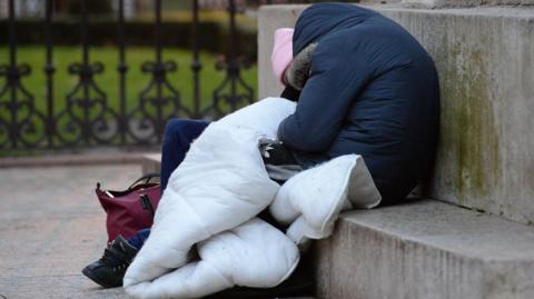 Person sleeping outside. They are wearing a navy coat with a fur lined hood and have a white bare duvet and a handbag.