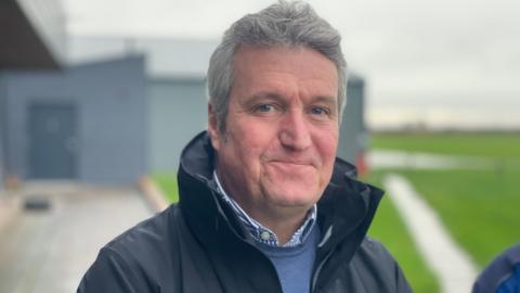 A man with short grey hair and wearing a blue rain coat is standing in front of buildings and looking into the camera. In the background are flooded areas of land.