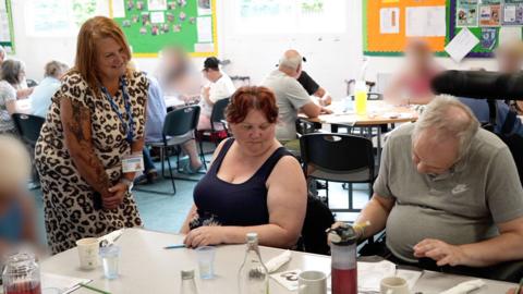 A woman wearing a leopard-print dress bends at a table where a man and woman sit. The man is filling out a form. There are mugs on the table with bottles of water. In the background more people sit around tables.
