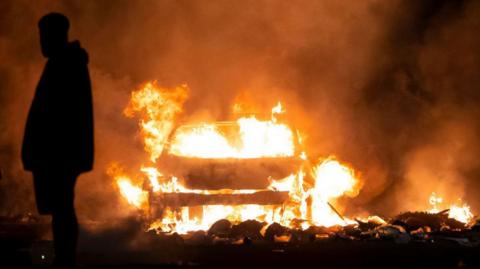 The silhouettes of a man shrouded in darkness with a large fire coming from a burning car in the background.