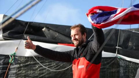 A bearded man in a sailing suit holds a union flag and in the other hand has a thumbs up.