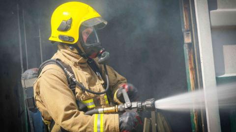 A firefighter in uniform and yellow hard hat holds a hose which is jetting out water