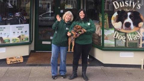 Two women wearing green hooded jumpers stand outside the front entrance of a shop. The woman on the right is holding a dog and a picture of a dog is visible on the shop window.