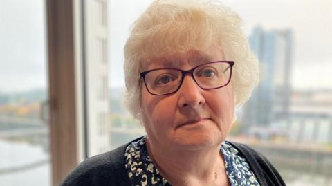 Laura Schosloff is staring at the camera. She has grey hair and glasses and is wearing a patterned black and white blouse and a black cardigan